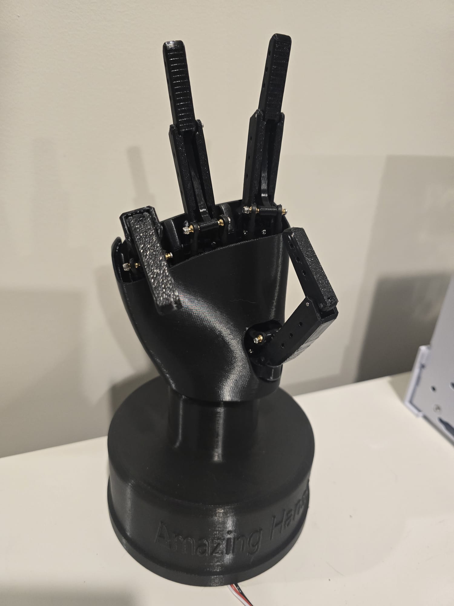 The black 3D-printed Pollen Robotics Amazing Hand robotic hand on a stand, fingers raised mid-gesture